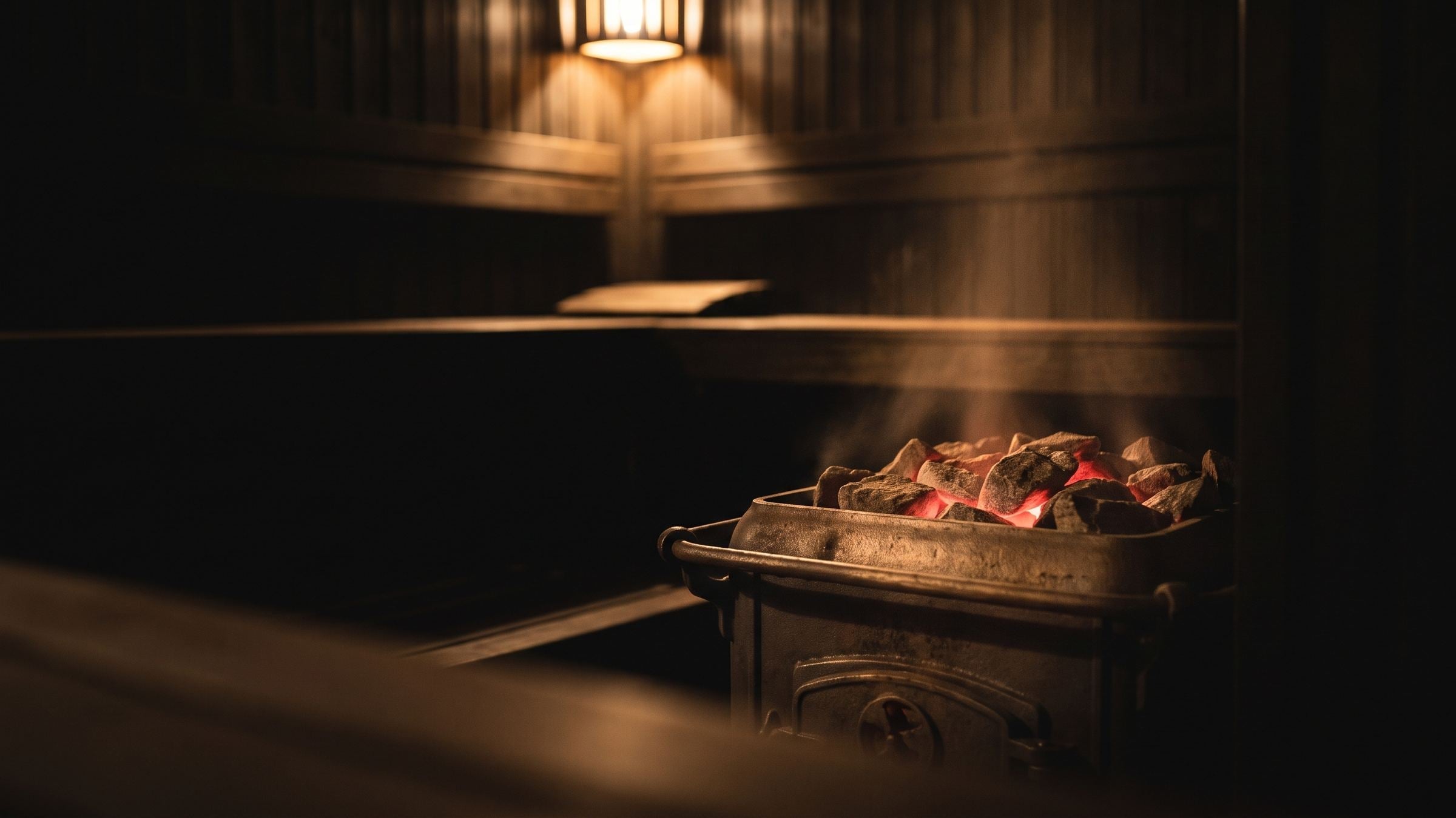 A sauna stove's coals glowing in a dark wood room, steam rising, a single pendant lamp overhead.