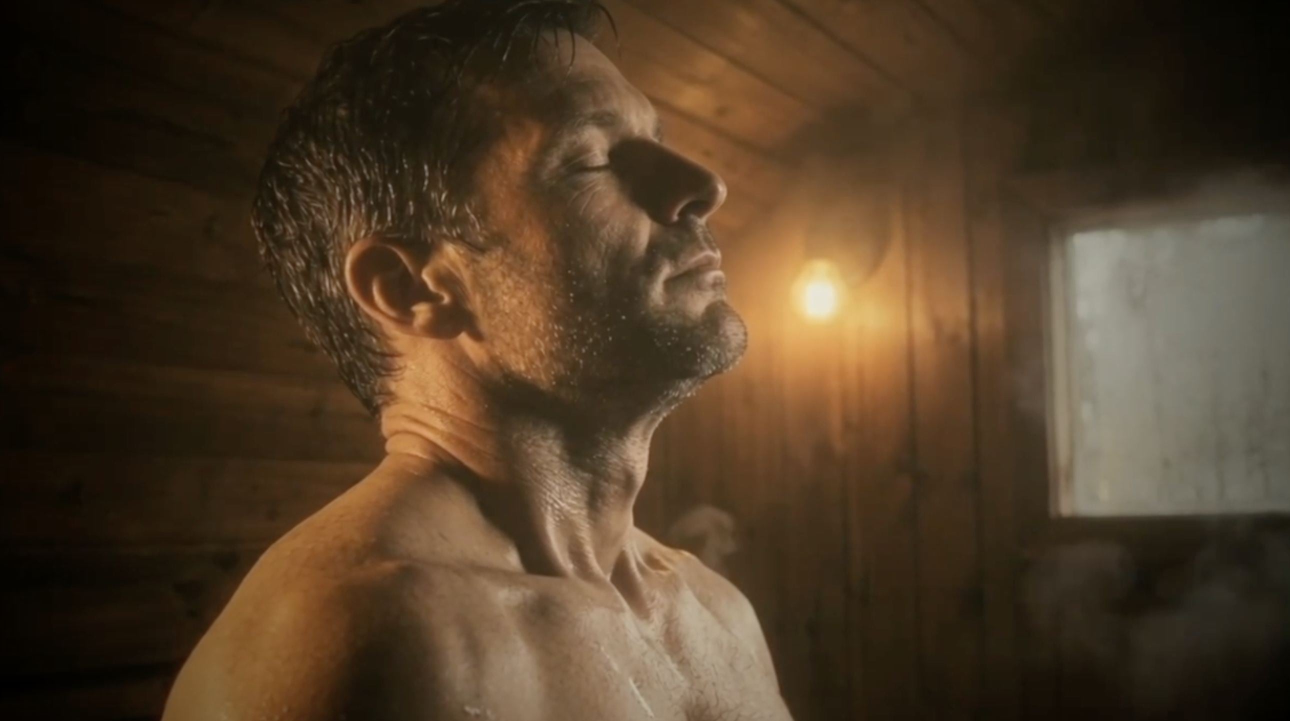A man in profile in a sauna, eyes closed, steam and window light behind him.